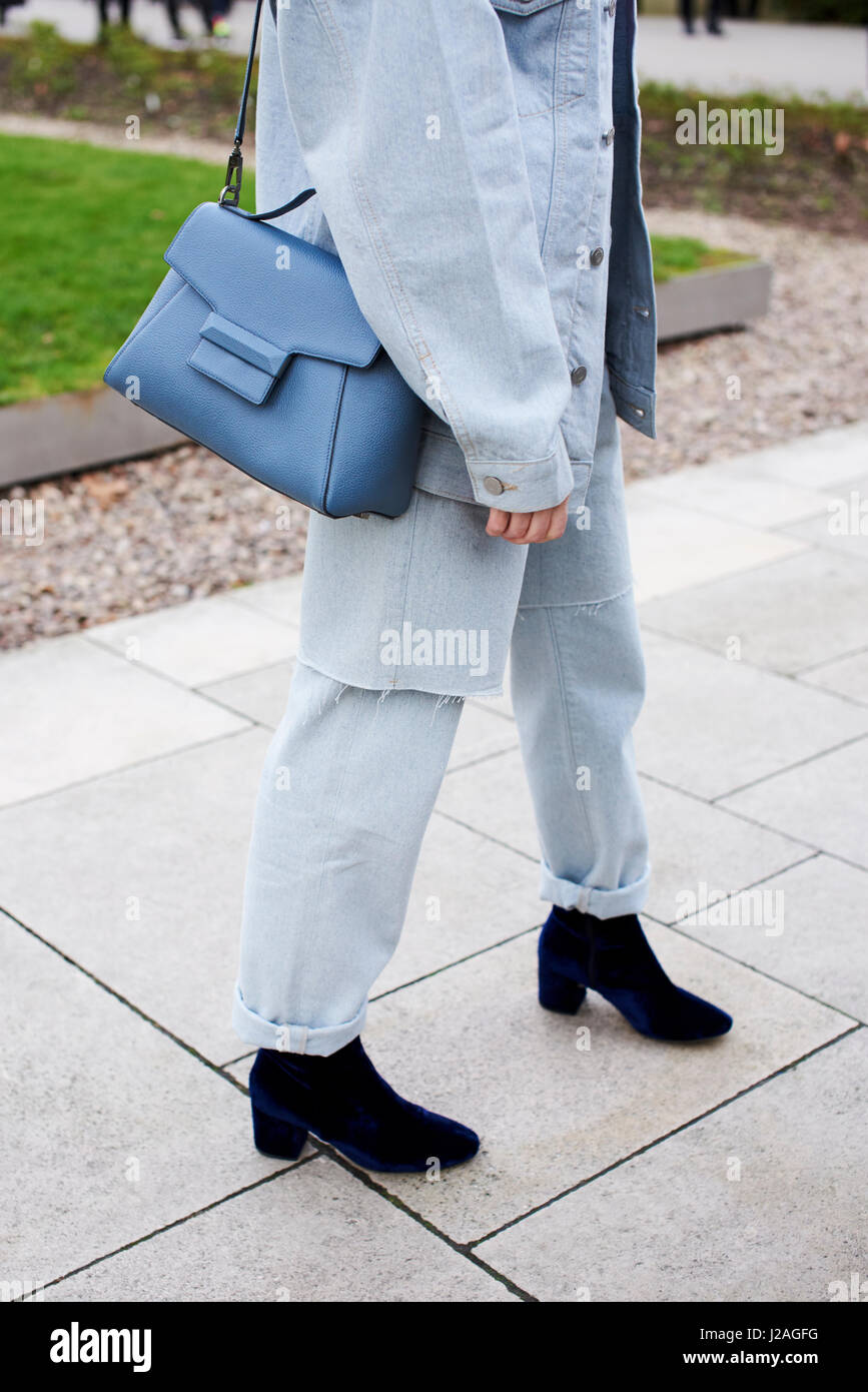 LONDON - FEBRUARY, 2017: Low section of woman wearing grey denim jacket and jeans with a blue leather shoulder bag turning towards camera in the street during London Fashion Week, vertical, side view Stock Photo