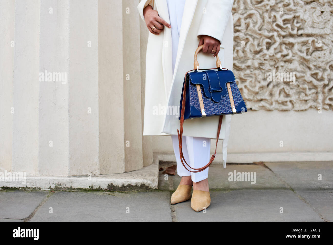 LONDON - FEBRUARY, 2017: Low section of woman wearing white trousers and coat holding a blue handbag standing in the street against a building during London Fashion Week, horizontal, front view Stock Photo