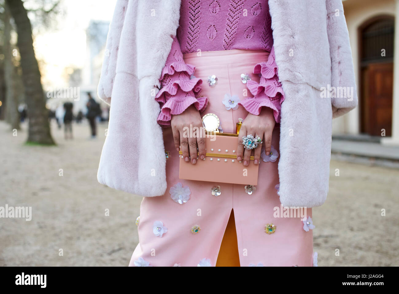 LONDON - FEBRUARY, 2017: Mid section of woman dressed in shades of pink, with a ruffle sleeved sweater, a coat over her shoulders and a skirt with appliqué, holding a decorated purse in the street during London Fashion Week, horizontal, front view Stock Photo