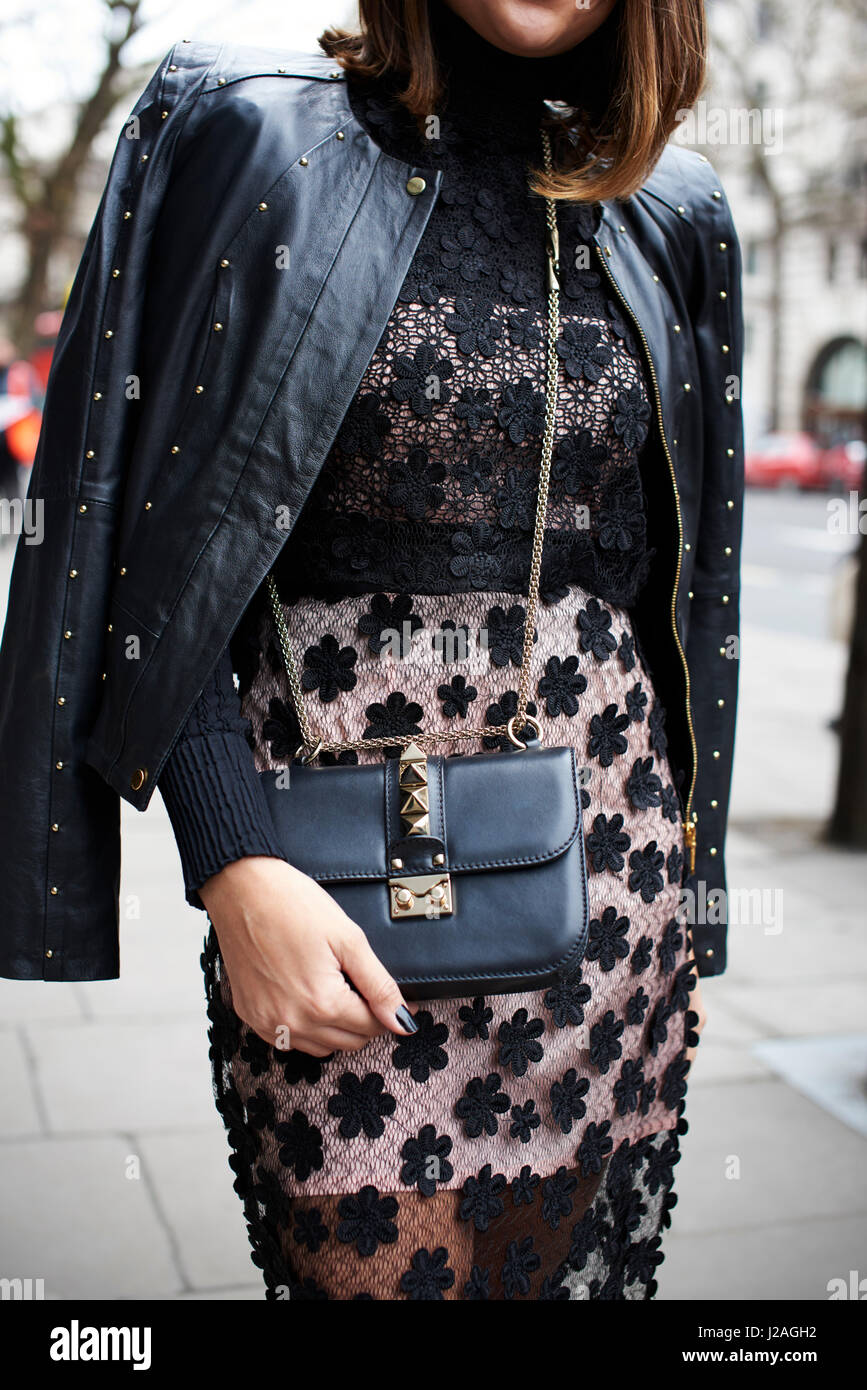 LONDON - FEBRUARY, 2017: Mid section of fashionable woman wearing a black lace dress with leather jacket over her shoulders and a Valentino cross body handbag, London Fashion Week, vertical, front view Stock Photo