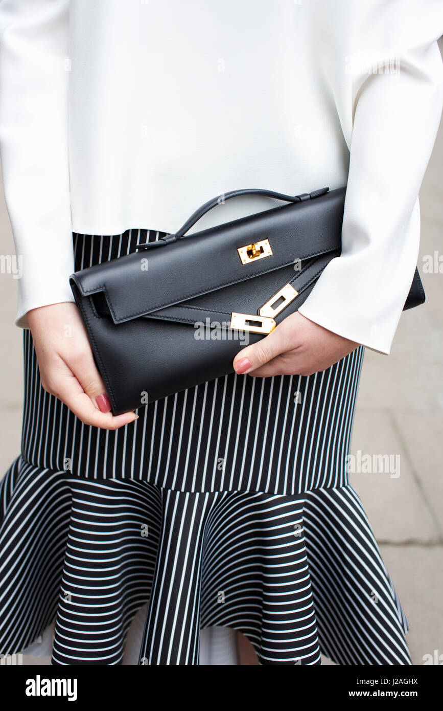 LONDON - FEBRUARY, 2017: Mid section of fashionable woman wearing a black and white striped skirt and white batwing top holding a Hermes clutch bag in the street during London Fashion Week, vertical, front view Stock Photo