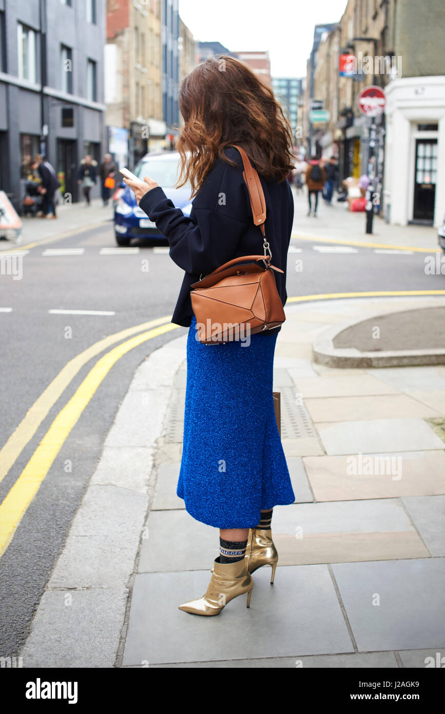 LONDON - FEBRUARY, 2017: Woman wearing blue wool skirt, socks and gold ankle boots standing in the street using phone during London Fashion Week, vertical Stock Photo