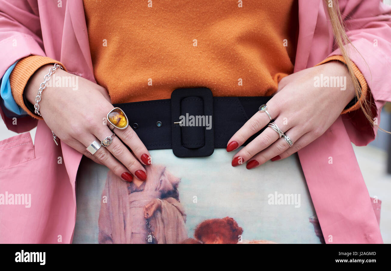 LONDON - FEBRUARY, 2017: Mid section detail of woman wearing pink coat and orange sweater with bold print skirt with black belt wearing rings in a street during London Fashion Week, horizontal Stock Photo