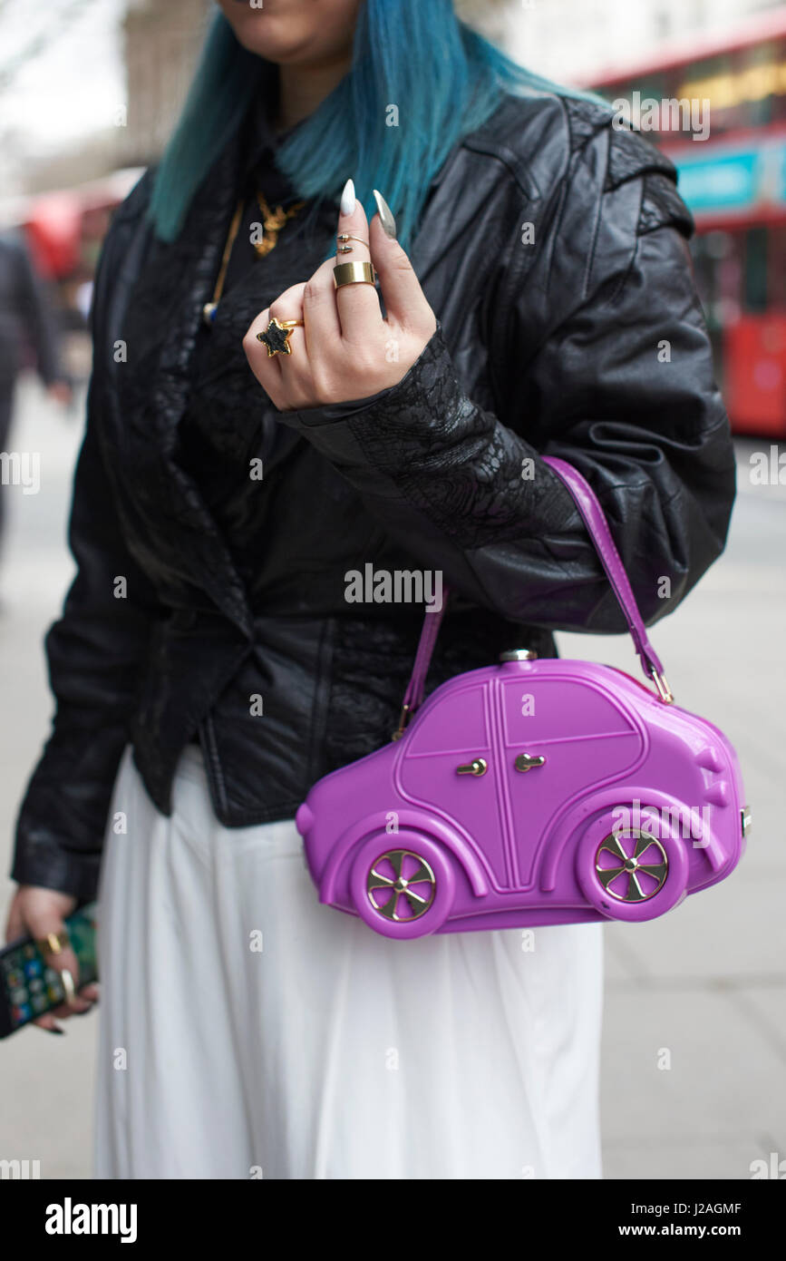 LONDON - FEBRUARY, 2017: Mid section of woman with blue hair wearing leather jacket and white trousers holding purple car-shaped handbag in a street during London Fashion Week, vertical Stock Photo