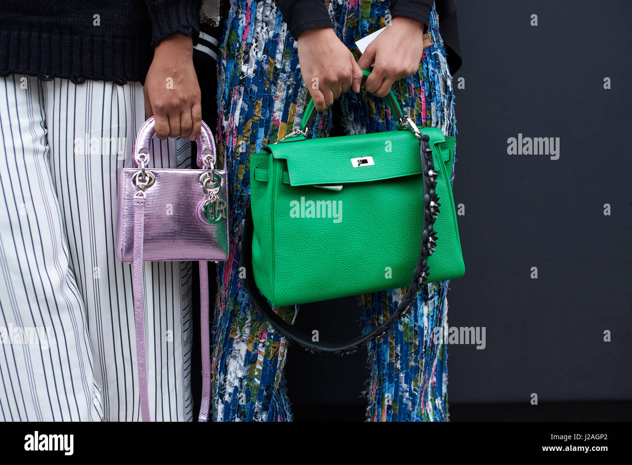 LONDON - FEBRUARY, 2017: Mid section of two women standing in a street holding designer handbags, a large green Hermes bag, and a small metallic Dior bag, during London Fashion Week, horizontal, front view Stock Photo