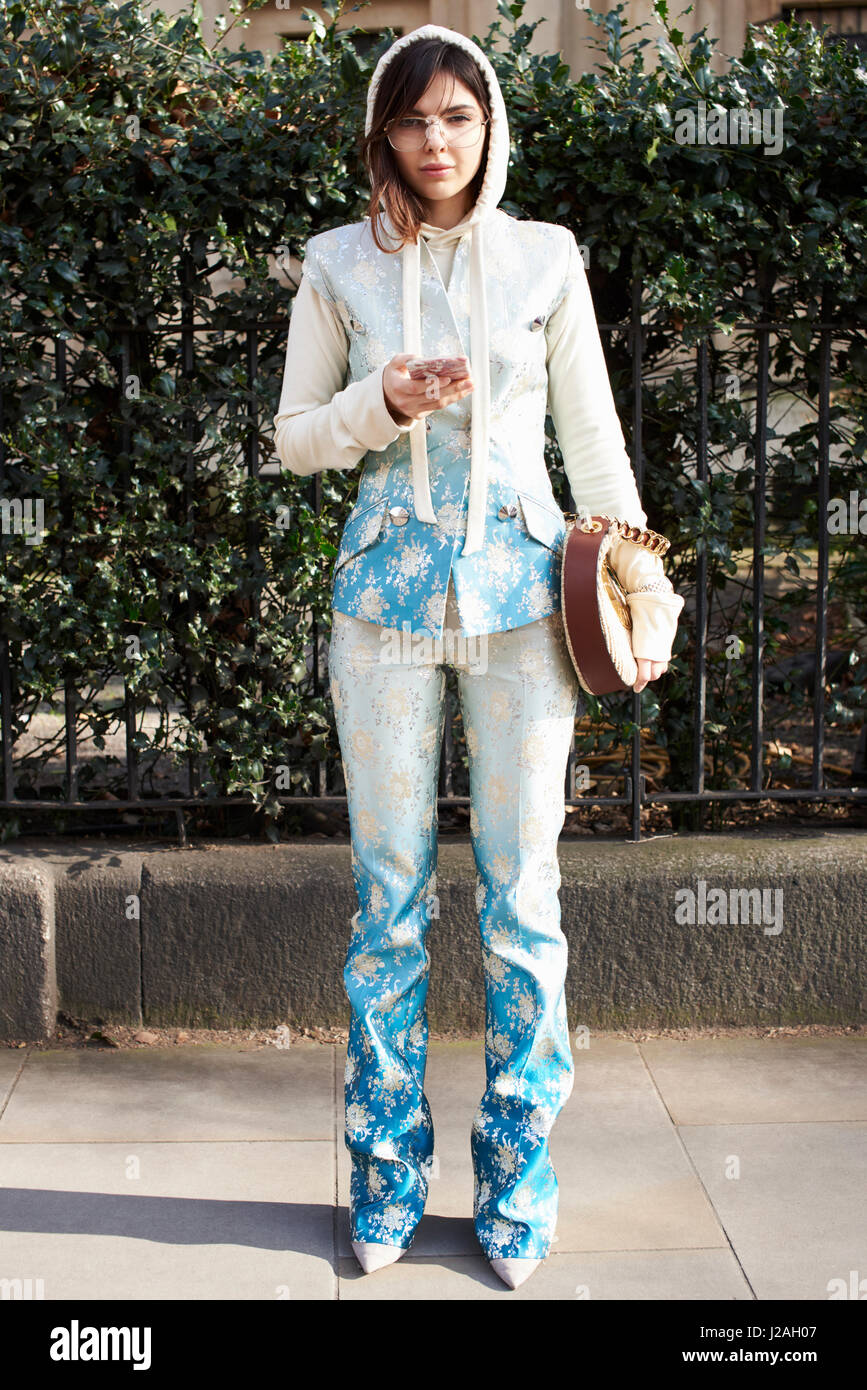 LONDON - FEBRUARY, 2017: Full length portrait of fashion model, stylist and blogger Doina Ciobanu wearing a blue and white patterned trouser suit with sleeveless jacket and white hoodie, holding a smartphone outside Pringle of Scotland show, London Fashio Stock Photo