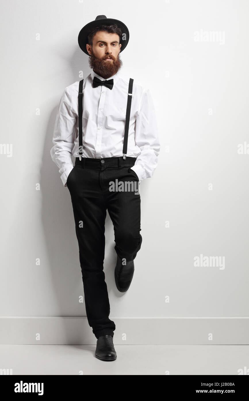 Full length portrait of an elegant man with suspenders and a bow tie leaning against a white wall Stock Photo