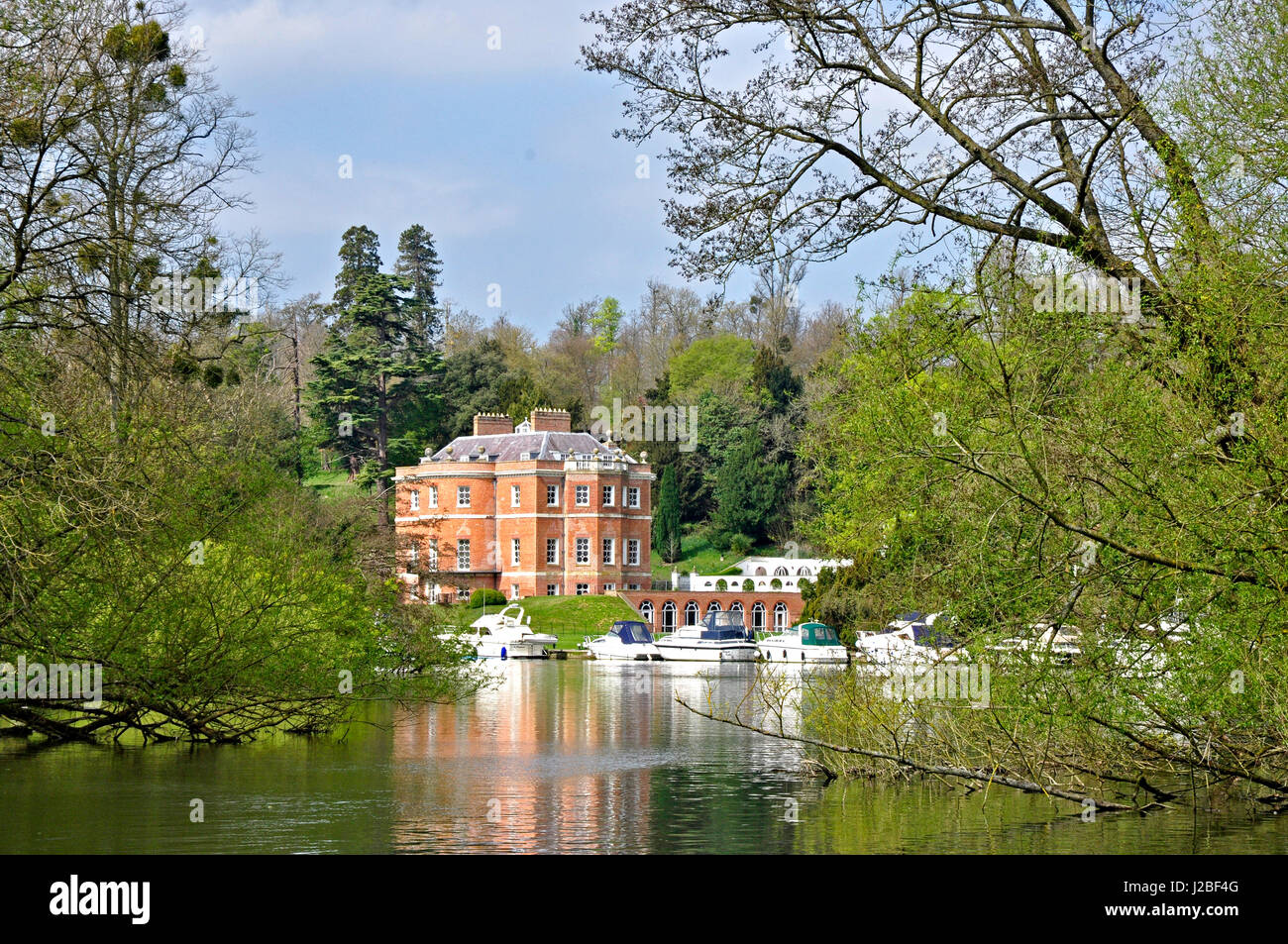 Harleyford Manor  Buckinghamshire across river Thames framed by springtime trees river cruisers moored  sunlight blue sky  reflections in the water Stock Photo