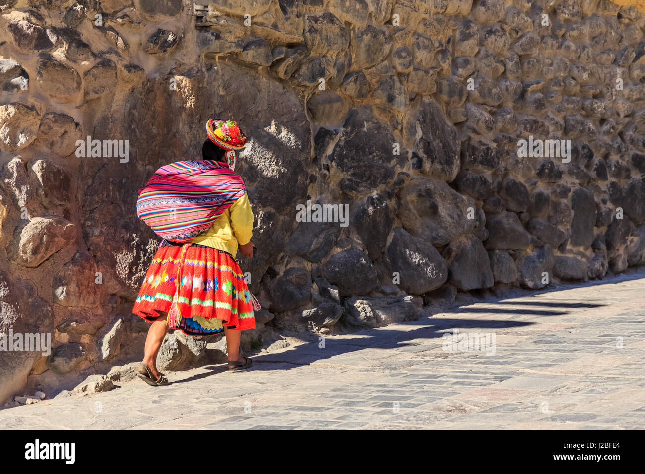 Peruvian woman cholita dressed in traditional colorful cloth, carrying the sack and walking up the street with stony walls, Inkan Sacred Valley, Ollan Stock Photo
