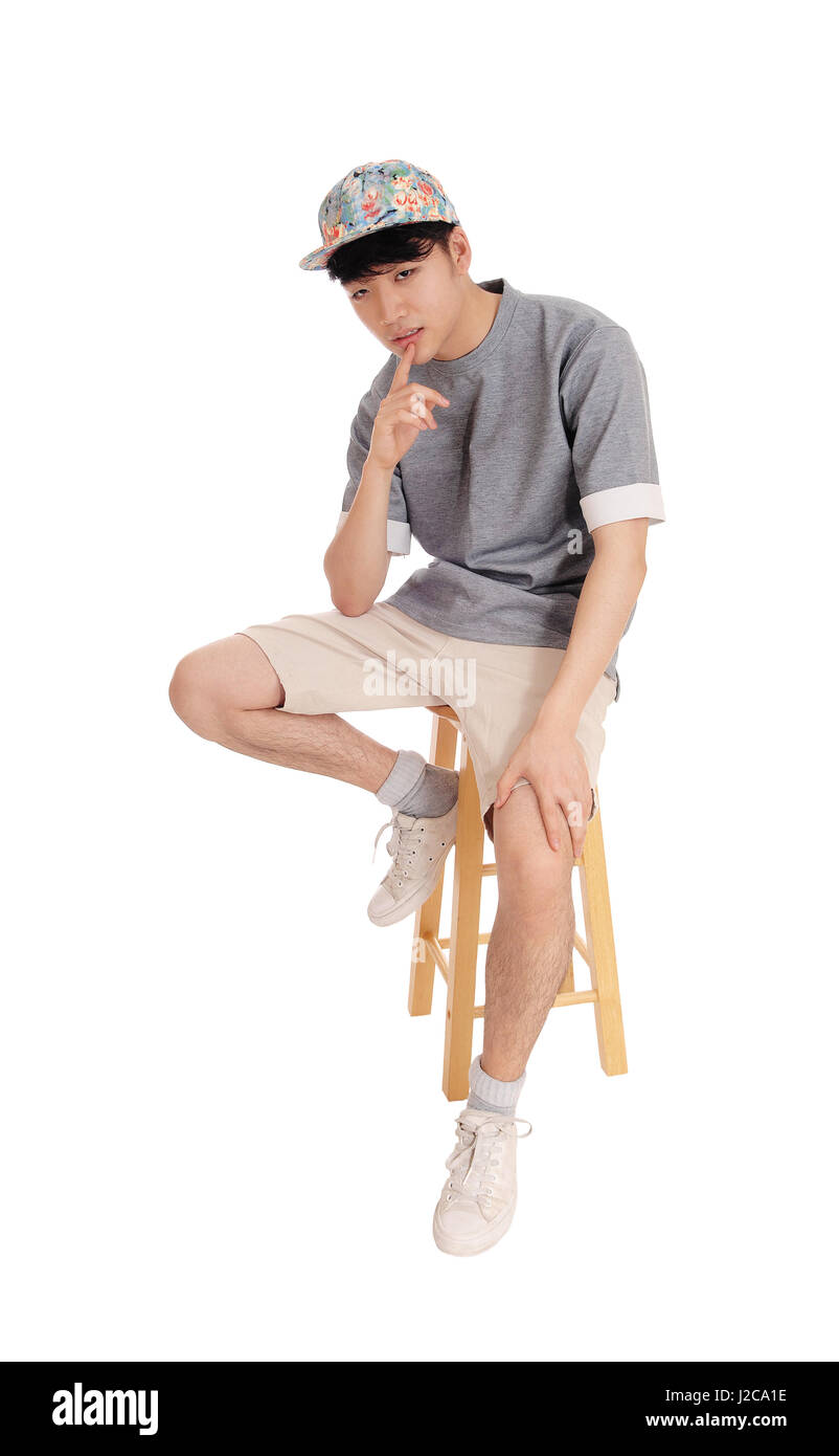 A young Korean teenager sitting on a chair in shorts and a cap with his finger on his chin, isolated for white background. Stock Photo
