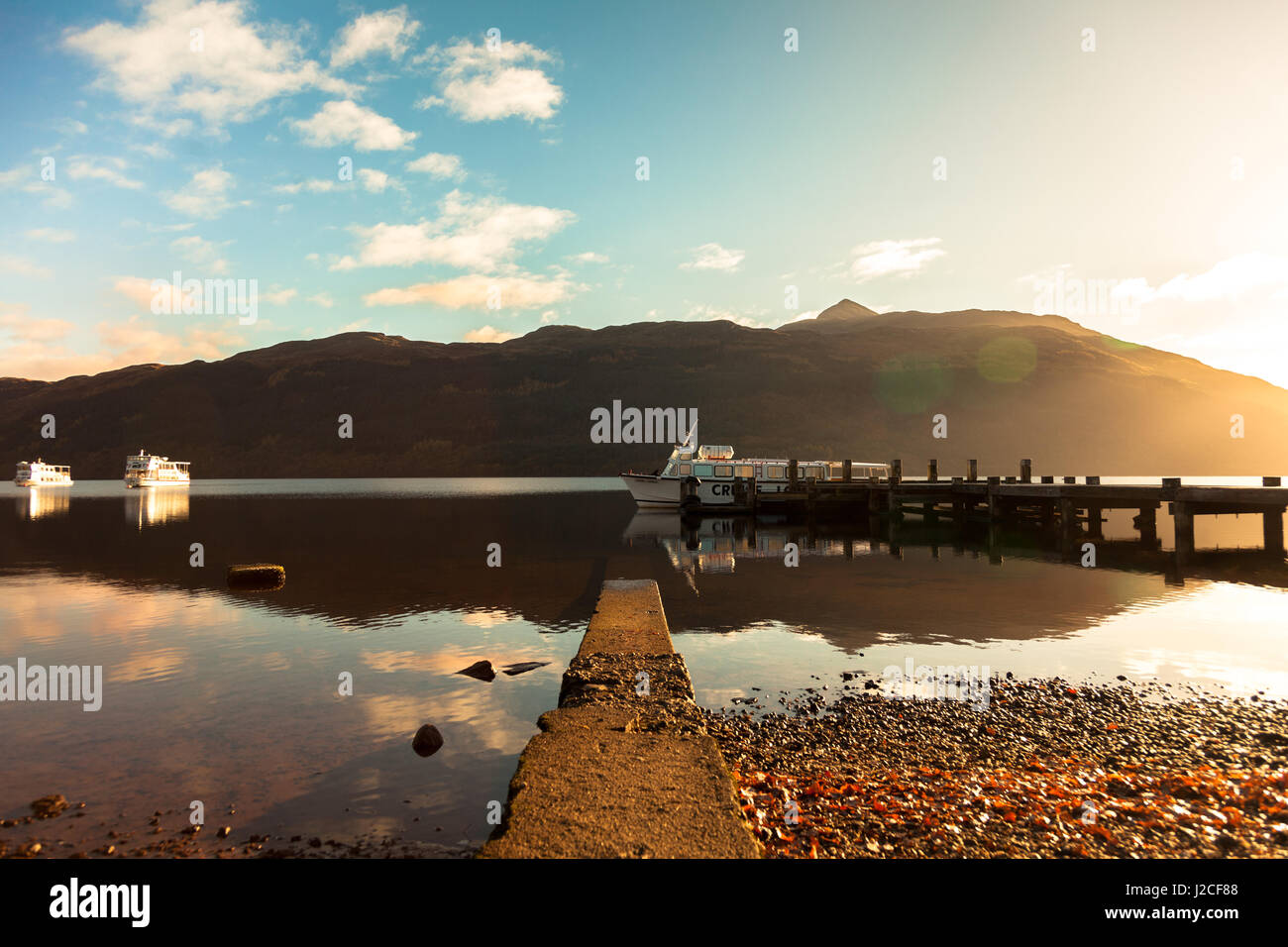 Sunset on the still mirror like waters of Loch Lomond as a boat floats at the end of a pier. Tarbet, Scottish Highlands, Scotland Stock Photo