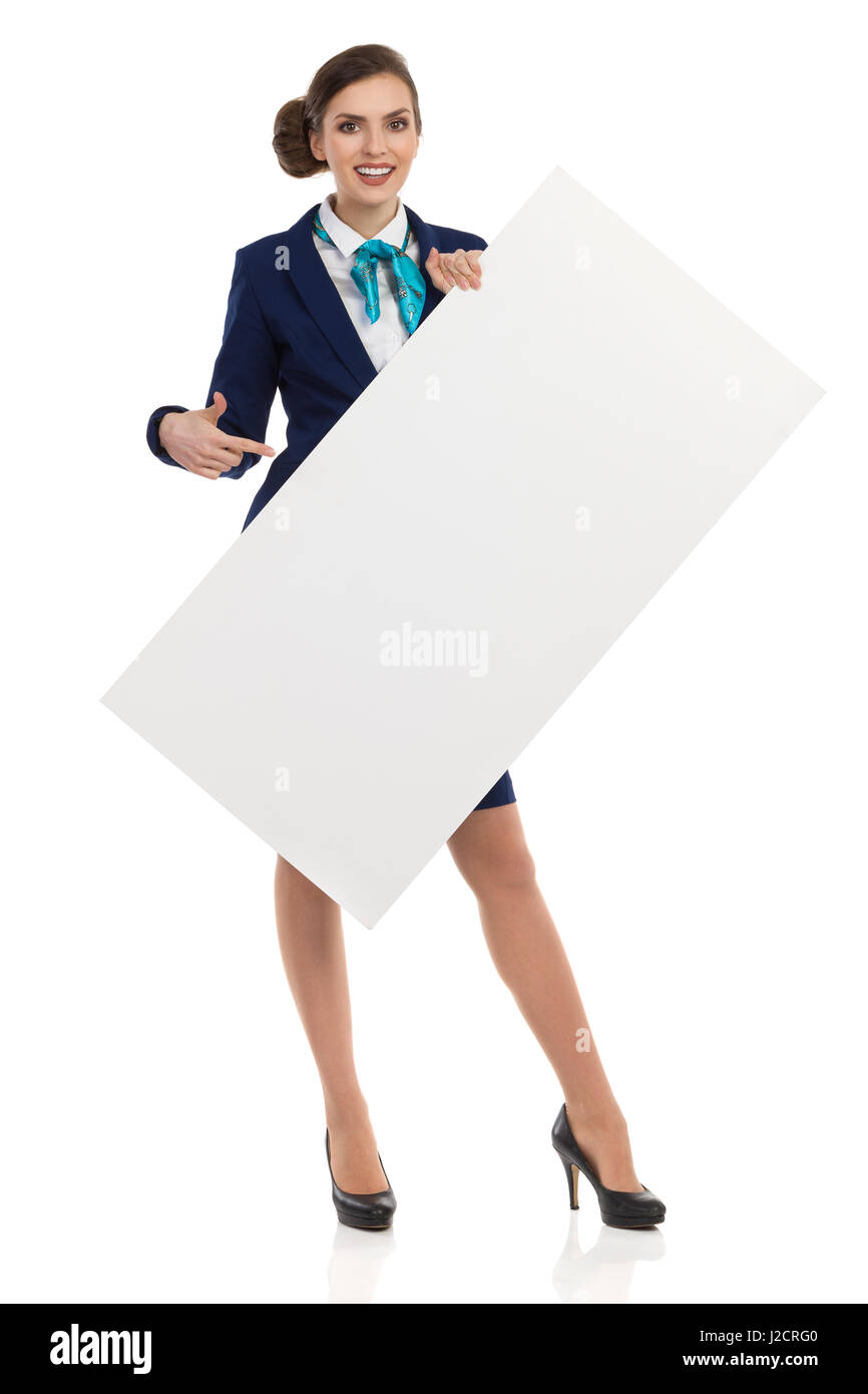 Young woman in blue formalwear and high heels, holding blank placard, pointing and smiling. Front view. Full length studio shot isolated on white. Stock Photo