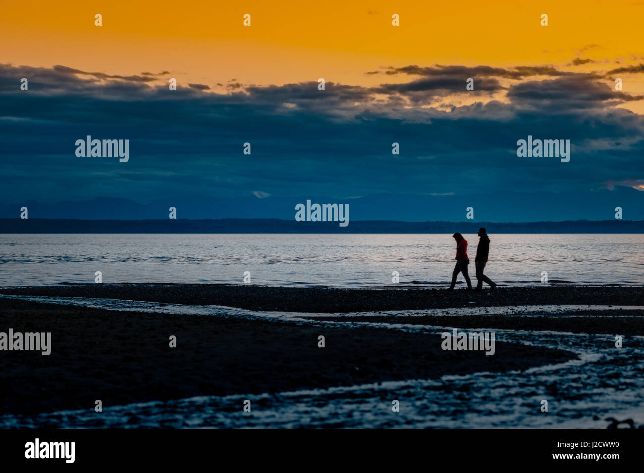 Couple strolling along the beach in a gloomy, cloudy sunset sky in Puget Sound Seattle. In walking, we find peace from the turmoils we are in in life. Stock Photo