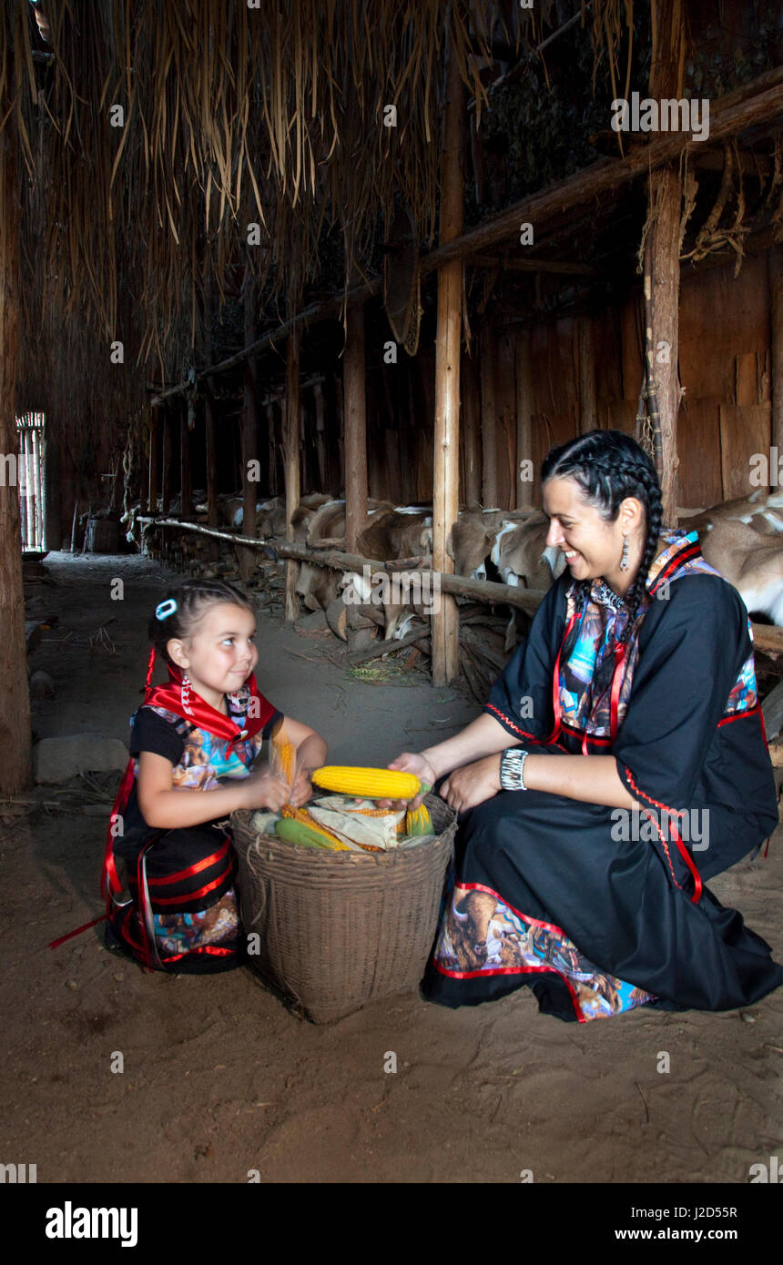 Ojibwe mother and daughter dressed in traditional ribbon dresses with a large basket of corn inside a bark covered longhouse (MR) Stock Photo