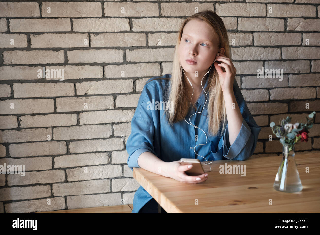 Beautiful casual blonde with a phone in her hand, touched the earpiece in the ear wearing in blue shirt at the cafe table. Copy space Stock Photo