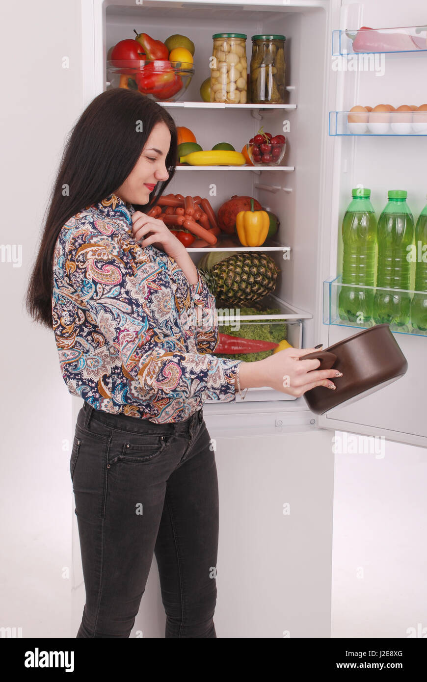Angry and upset housewife looking into a pot with foul meal. Beautiful young girl near the fridge. Stock Photo
