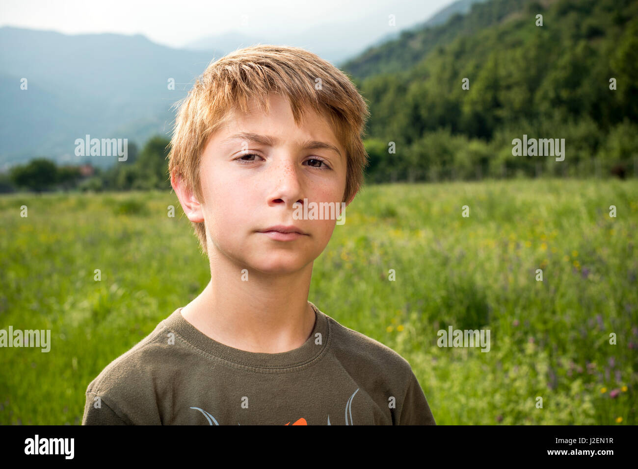 Head and Shoulders Portrait of Serious Young Boy Standing in Field with Mountains in Background Stock Photo