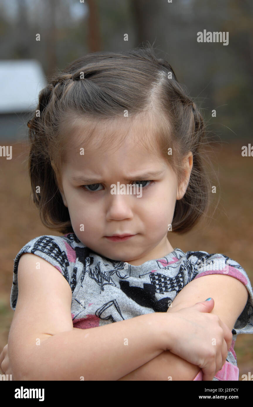 Arms crossed and eyebrows puckered, this little girl is upset and pouting.  She is standing outside and image is closeup. Stock Photo