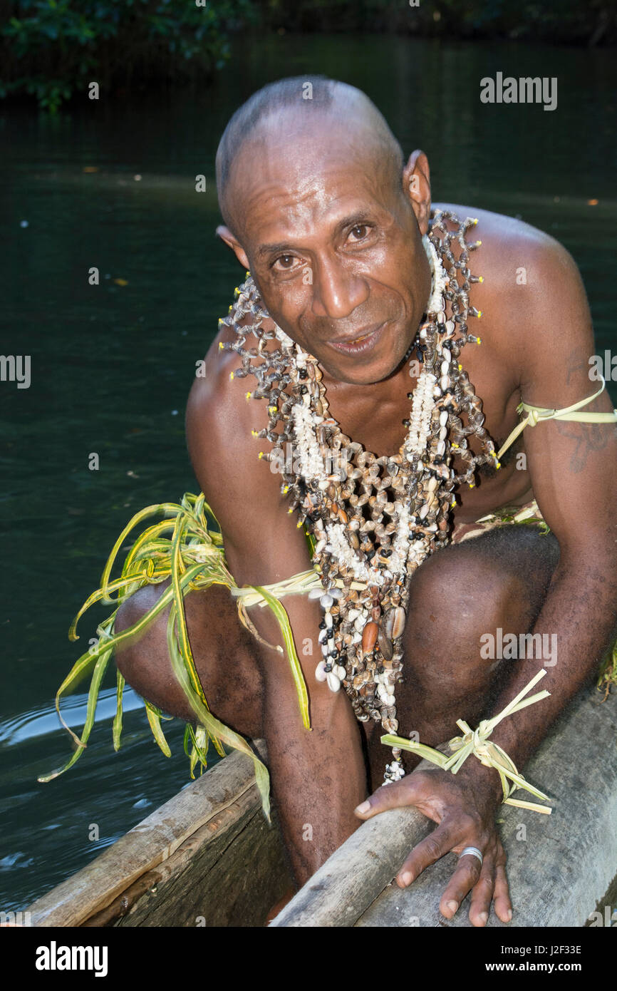 Melanesia, Papua New Guinea, Tufi. Village man in traditional attire with seashell necklaces paddling dugout canoe on remote river. Bailing water out of bottom of the canoe. Stock Photo