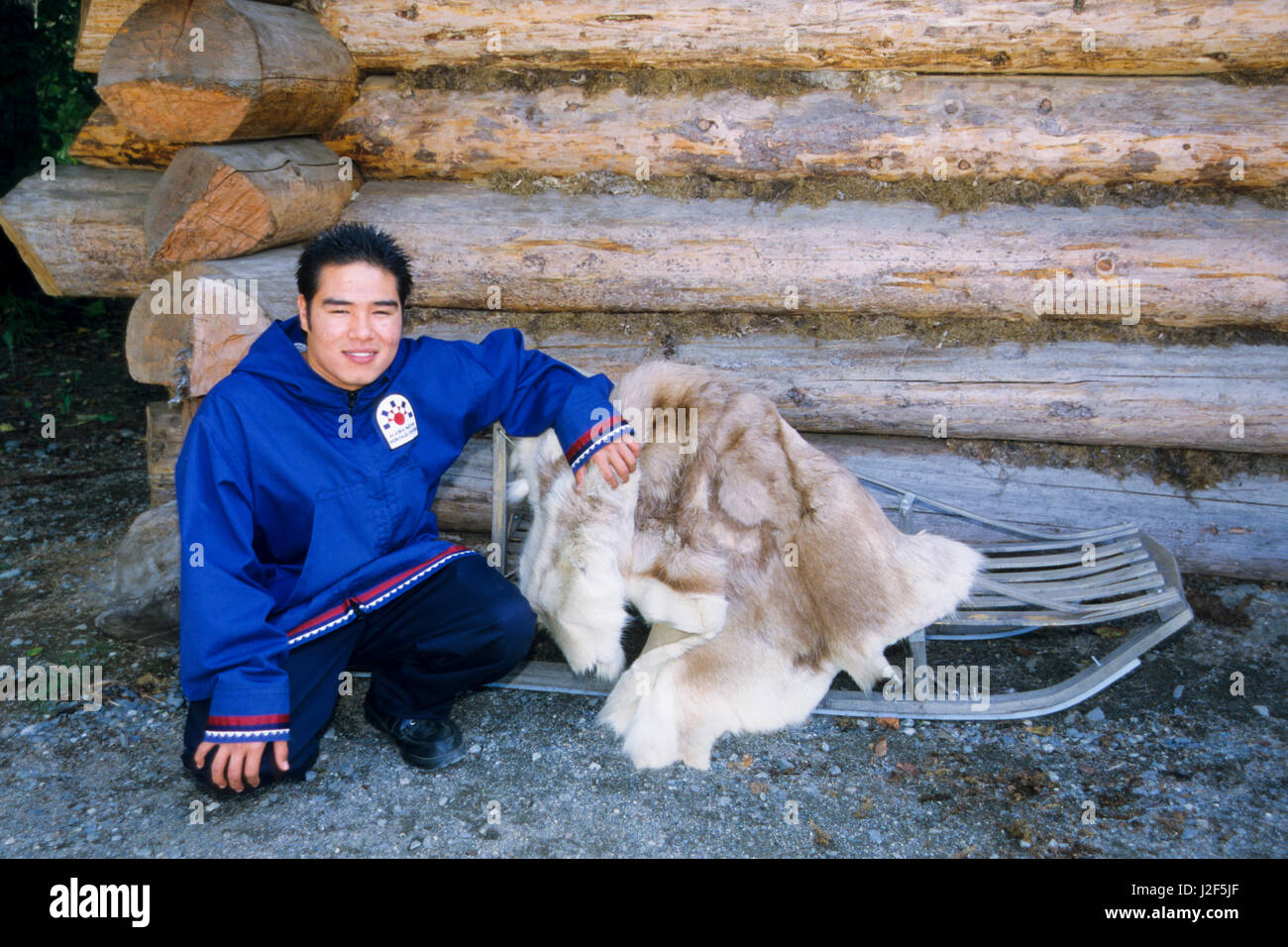 Native Interpreter next to wooden dog sled and caribou fur hide outside of Athabaskan log dwelling. Alaska Native Heritage Center, Anchorage Alaska. Stock Photo