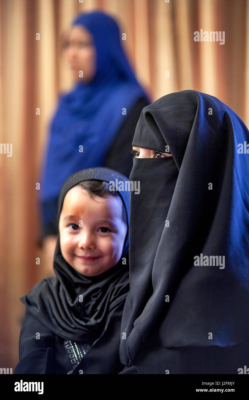A Muslim mother wearing a face-covering burqua holds her toddler son in traditional Middle Eastern clothing at an Anaheim, CA, mosque. Note woman in background wearing a hijab, the traditional Muslim female head scarf. Stock Photo