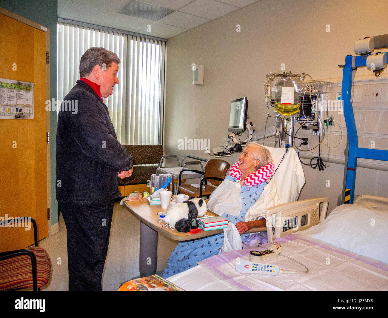 A Catholic priest pays an encouraging visit to a senior woman patient in bed at an Irvine, CA, hospital. Note cervical collar. Stock Photo