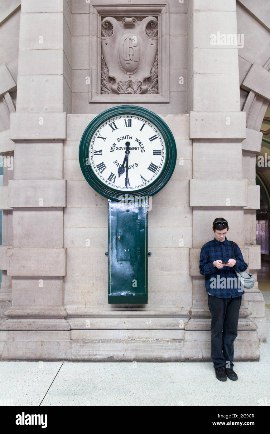 Young man checking his mobile phone by the side of a large clock in Central Railway Station, Sydney, Australia Stock Photo