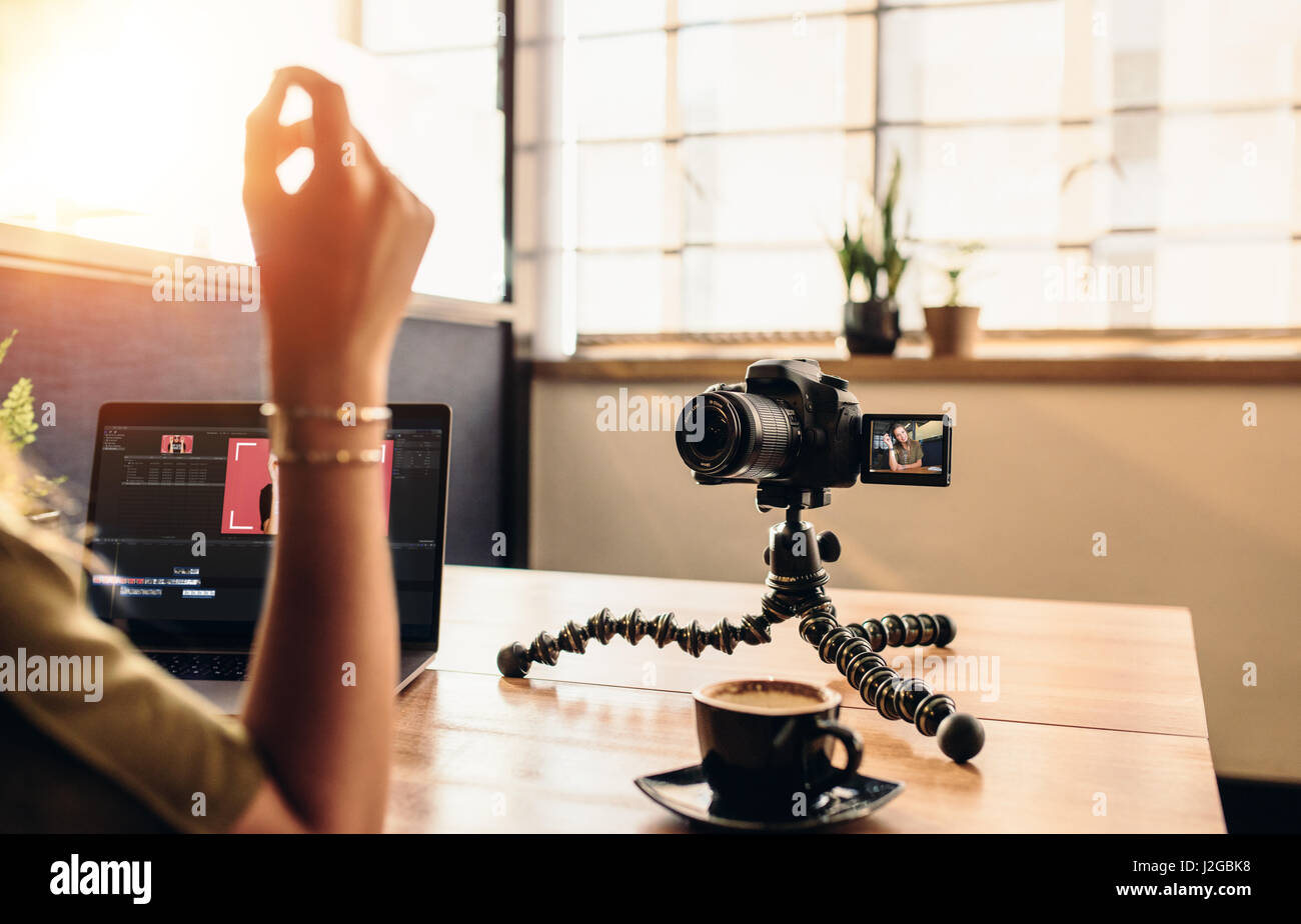 Female vlogger looking at camera while editing video on her laptop. Young woman working on computer with coffee and camera on her desk. Stock Photo