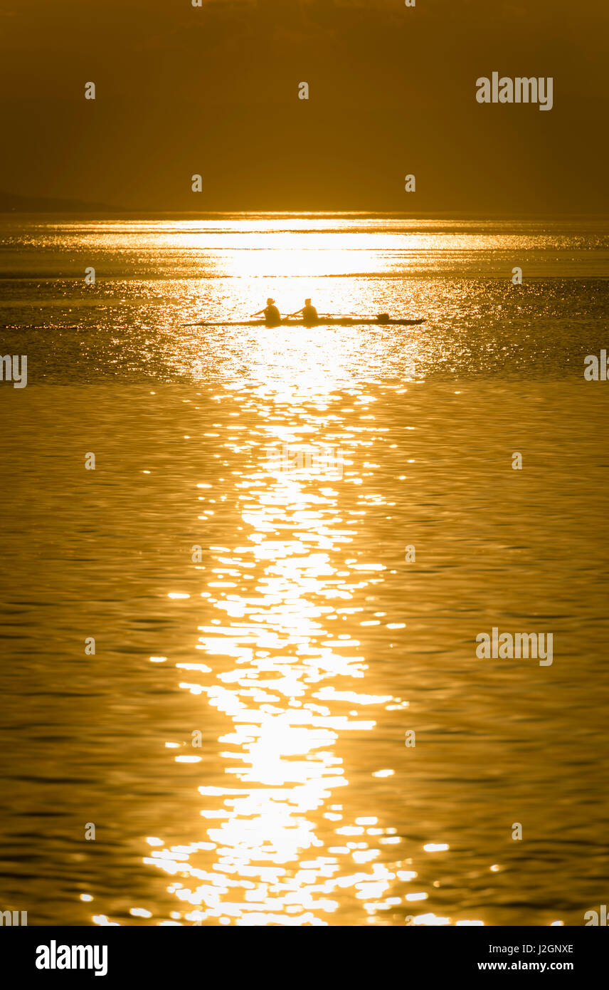 A team of two oarswomen is training in their rowboat on Lake Geneva, Switzerland. Back-lit, evening sunset. Stock Photo