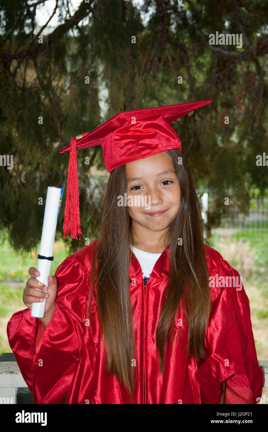 Native American elementary age girl dressed in graduation cap and gown holds up her diploma for passing her grade. Stock Photo