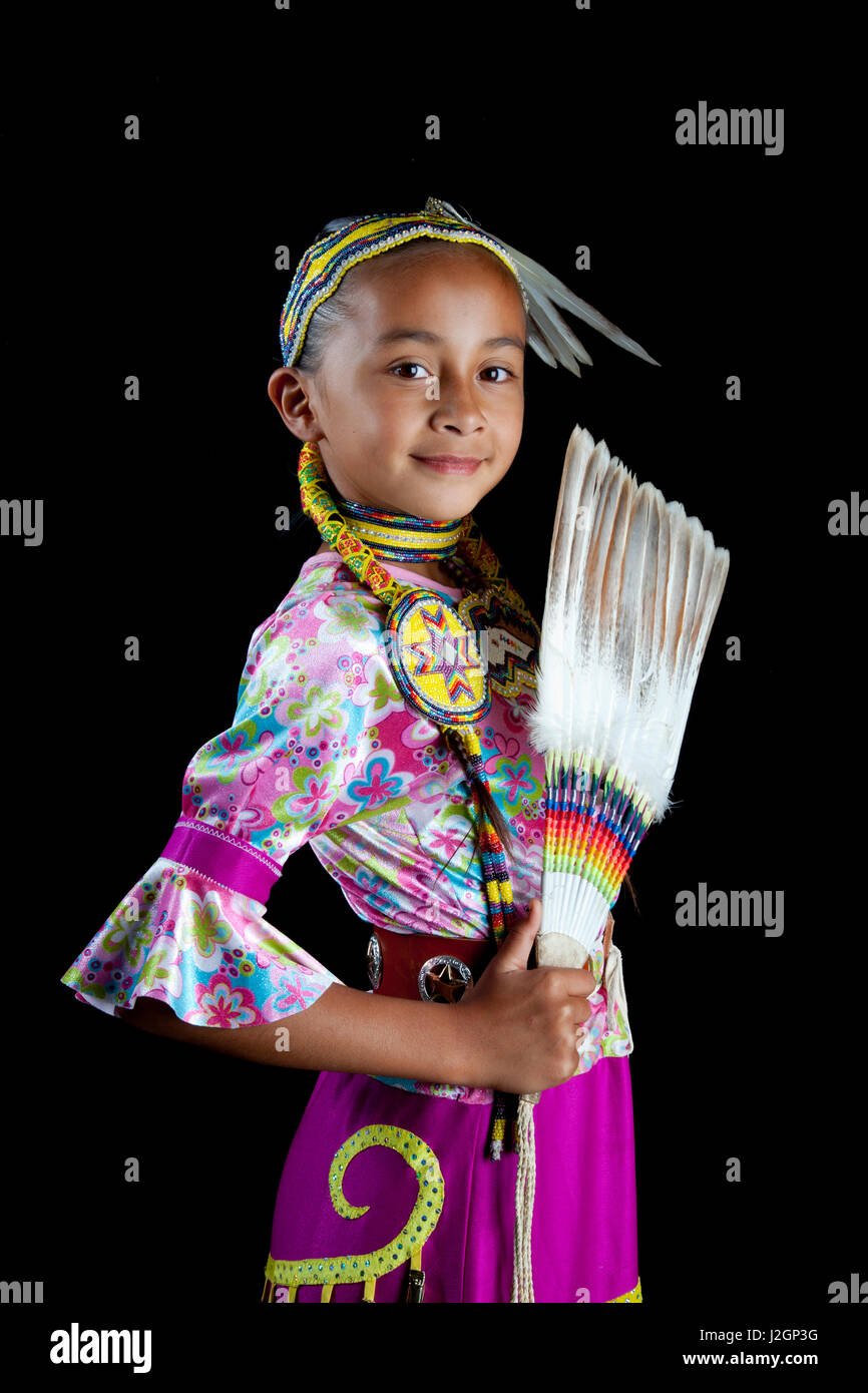 Traditional pow wow dancer 9 year old Jaydean (Lakota) dressed in colorful regalia and beaded hair band holds up a feather fan against a black background. Stock Photo