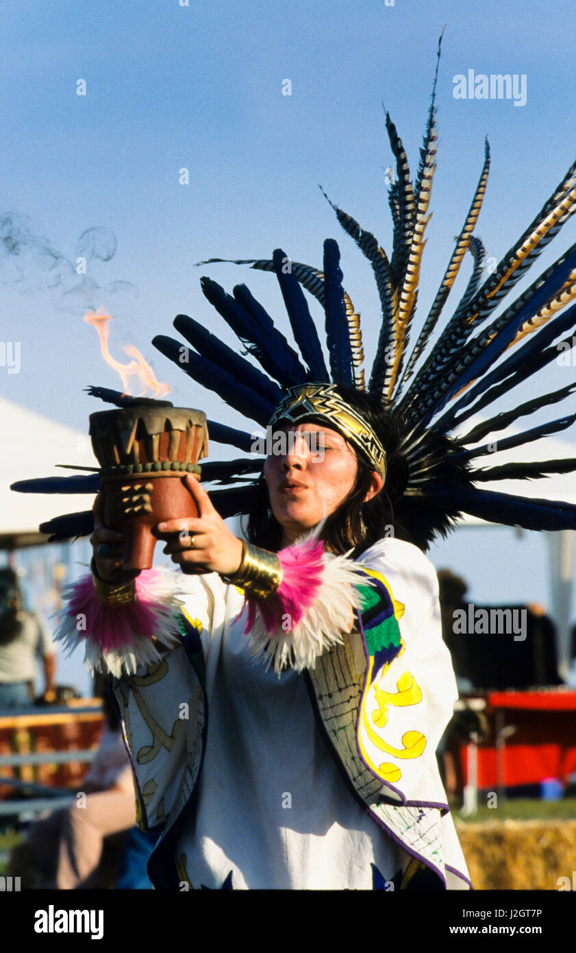 Aztec woman dressed in traditional dance regalia and feather headdress holds up a bowl with fire burning a medicine bundle. Stock Photo