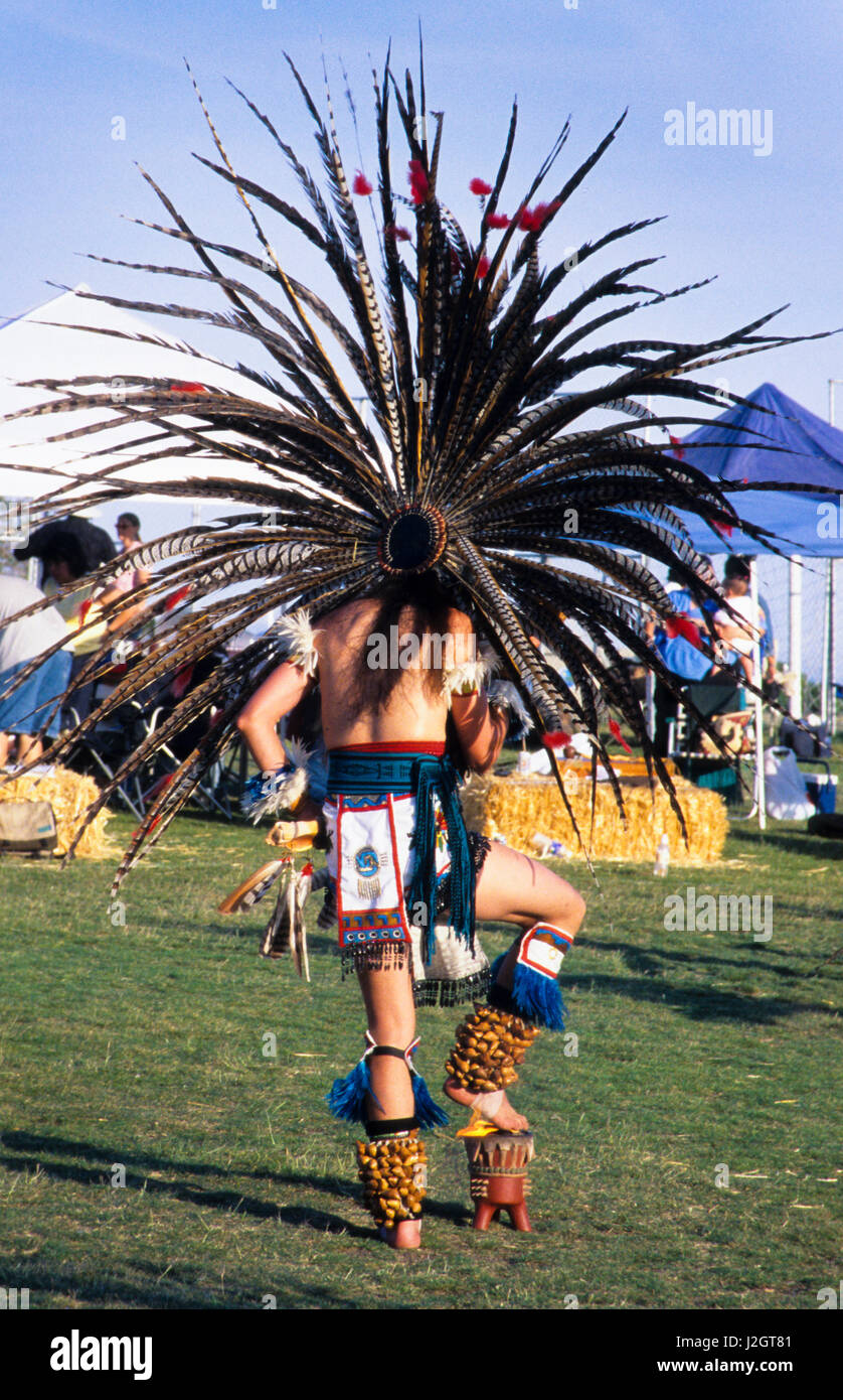 Aztec man dressed in regalia and large traditional feather headdress holds up a gourd rattle during a fire dance where he places his foot over the flame. Stock Photo