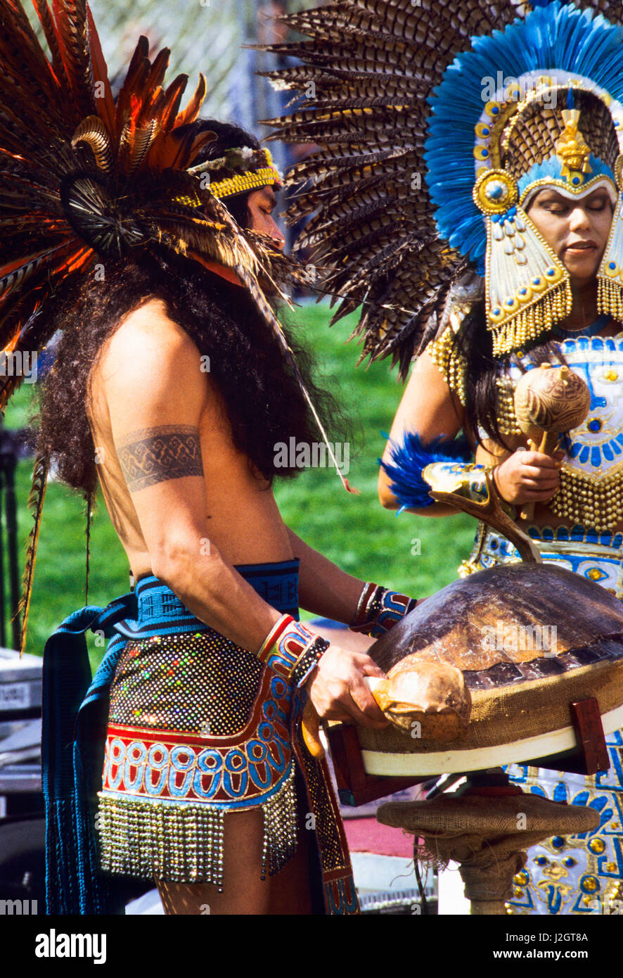 Aztec man dressed in traditional regalia plays a large turtle shell drum and a woman also dressed in ceremonial clothing and large feather headdress shakes a gourd rattle during a celebration of Aztec songs and dance. Stock Photo