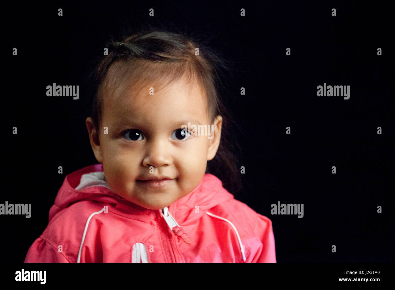 Contemporary Native American girl at toddler age in pink jacket next to black backdrop. Harmony Lahr (Blackfoot, Navajo) Stock Photo