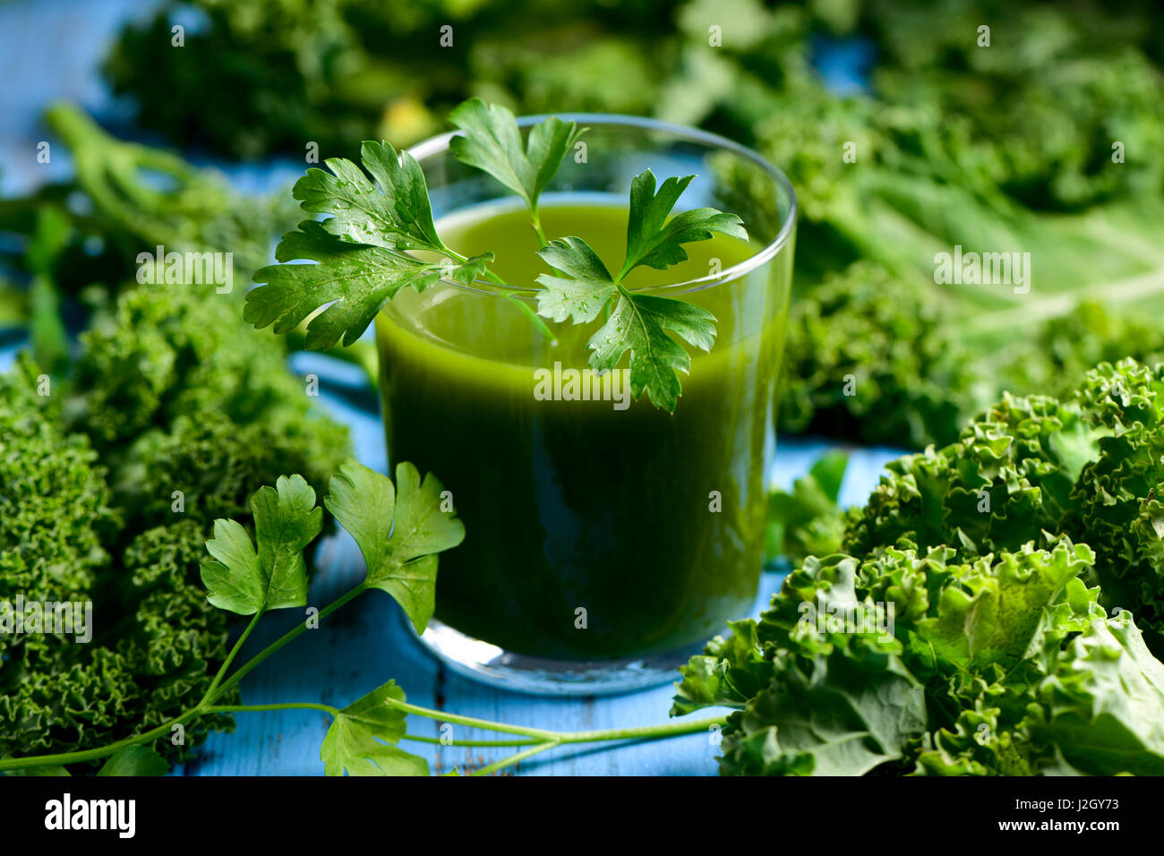 a glass with kale smoothie topped with a twig of parsley and some leaves of kale on a blue rustic wooden table Stock Photo
