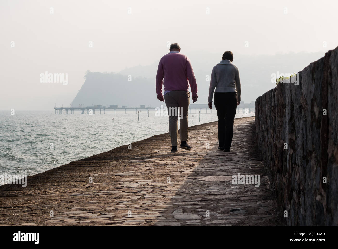 A man and a women walk along Teignmouth sea front railway path with the pier and The Ness rock in the background. Stock Photo