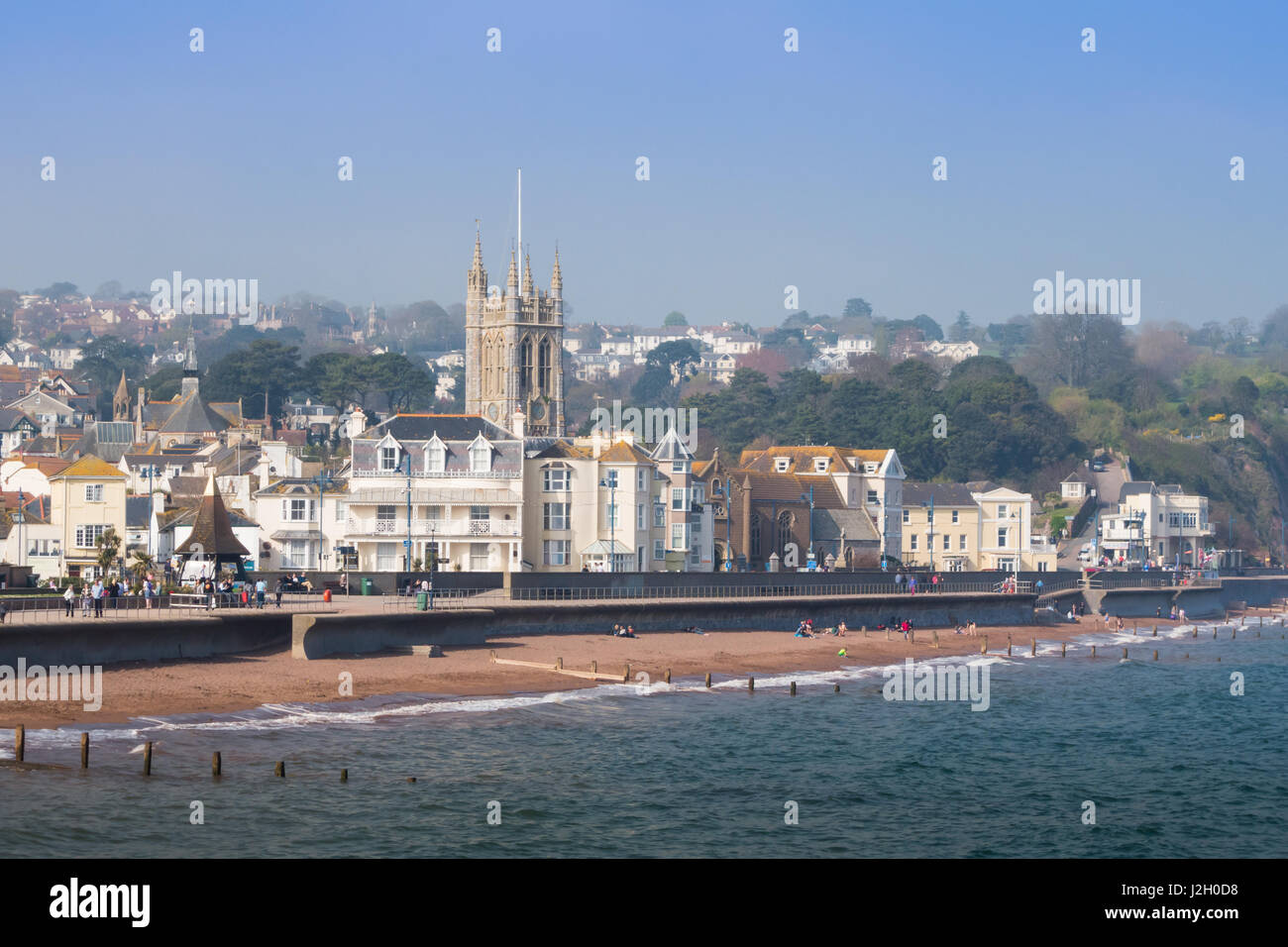 Views of Teignmouth sea front on a sunny Spring day with people walking along the promenade. St Michaels church is prominent in the background. Stock Photo