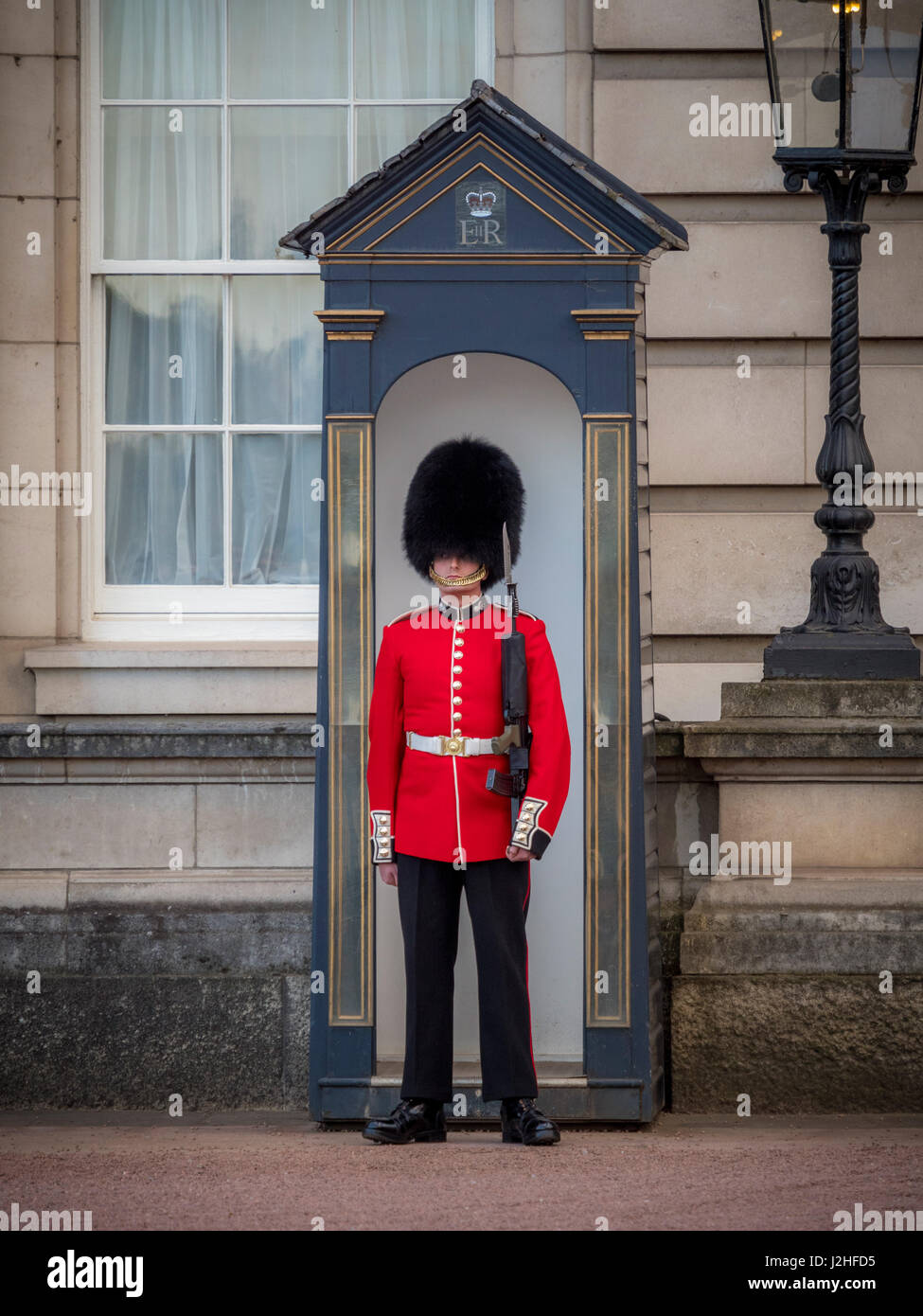 The grand facade of Buckingham Palace in London, with a Queen's Guard in traditional uniform standing sentry in front of the ornate gates.
