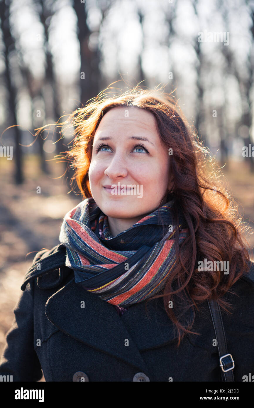 Smiling red-haired girl outdoors, weared coat and scarf. Early spring in park ar sunset. Stock Photo