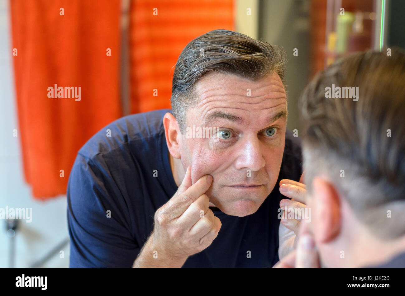 Middle aged caucasian man stands at mirror and pinches his face while wearing a blue shirt Stock Photo