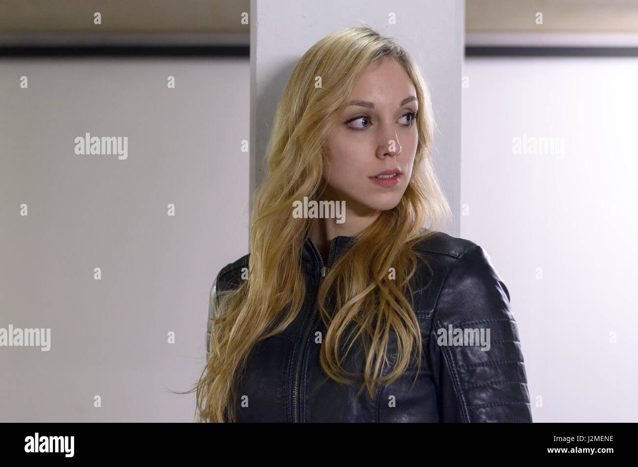 Attractive serious young blond woman standing leaning against a pillar looking to the side with an intent expression as she observes something off fra Stock Photo