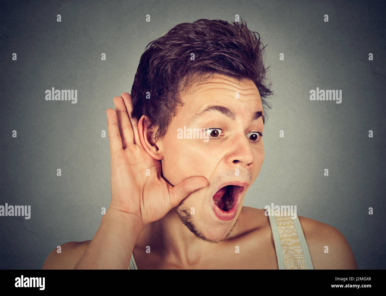 Closeup portrait surprised young nosy man hand to ear gesture carefully secretly listening gossip conversation news isolated grey background. Human fa Stock Photo