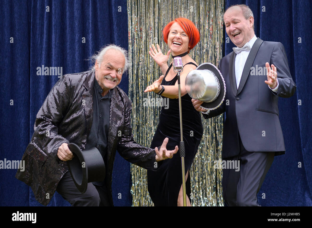 Three lively performers on stage celebrating in front of a microphone with a vivacious redhead woman and two gentlemen with top hats laughing and joki Stock Photo