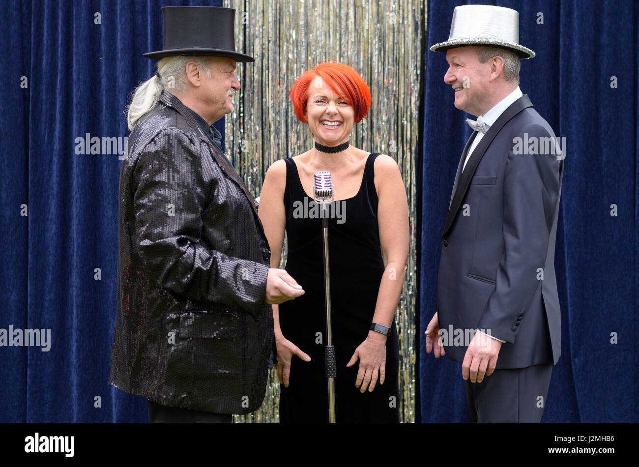 Three lively performers on stage celebrating in front of a microphone with a vivacious redhead woman and two gentlemen with top hats laughing and joki Stock Photo