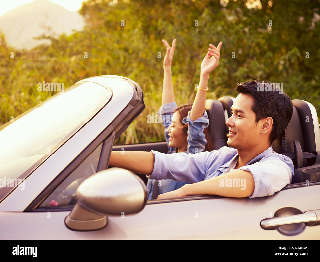 young asian couple riding in a convertible sport car enjoying the cool breeze at sunset. Stock Photo
