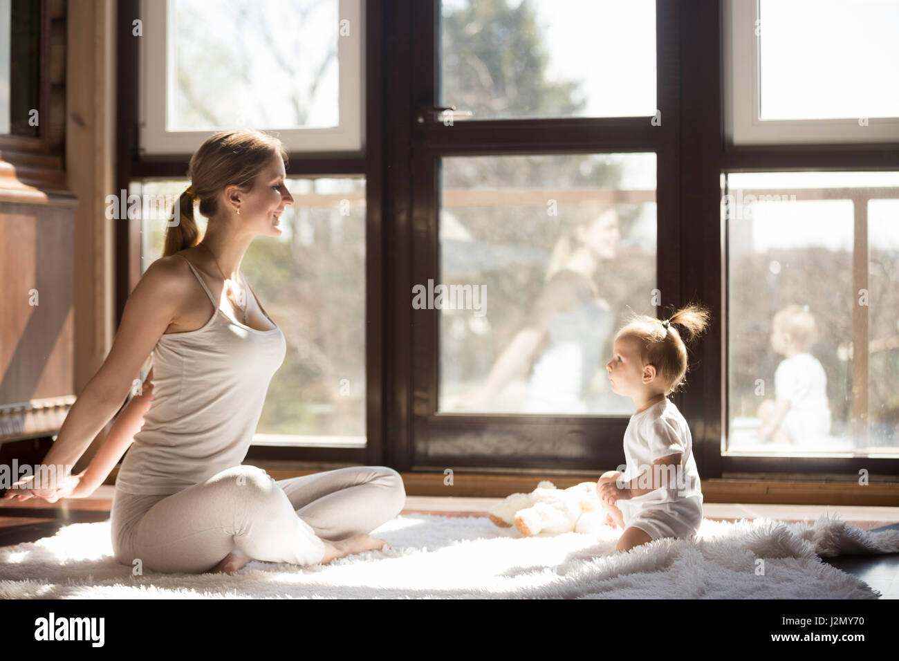 Young yogi mother at Easy Seat pose, cute baby daughter sitting near her, starting a new sporty day together, enjoying morning exercises at home, fami Stock Photo