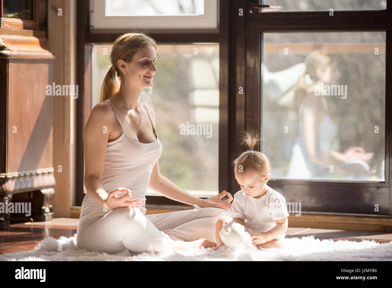 Young yogi mother in Easy Seat pose, wearing white sportswear, baby daughter playing near her, starting a new day in a good mood, harmony and relaxati Stock Photo