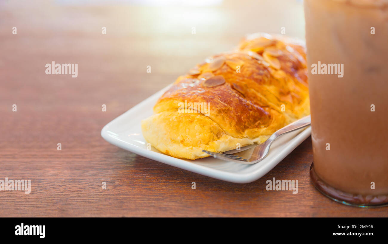 Almond croissant breakfast on a white plate and fork, serves with cold chocolate drink, all on wooden table, selective focus on the front part of the  Stock Photo