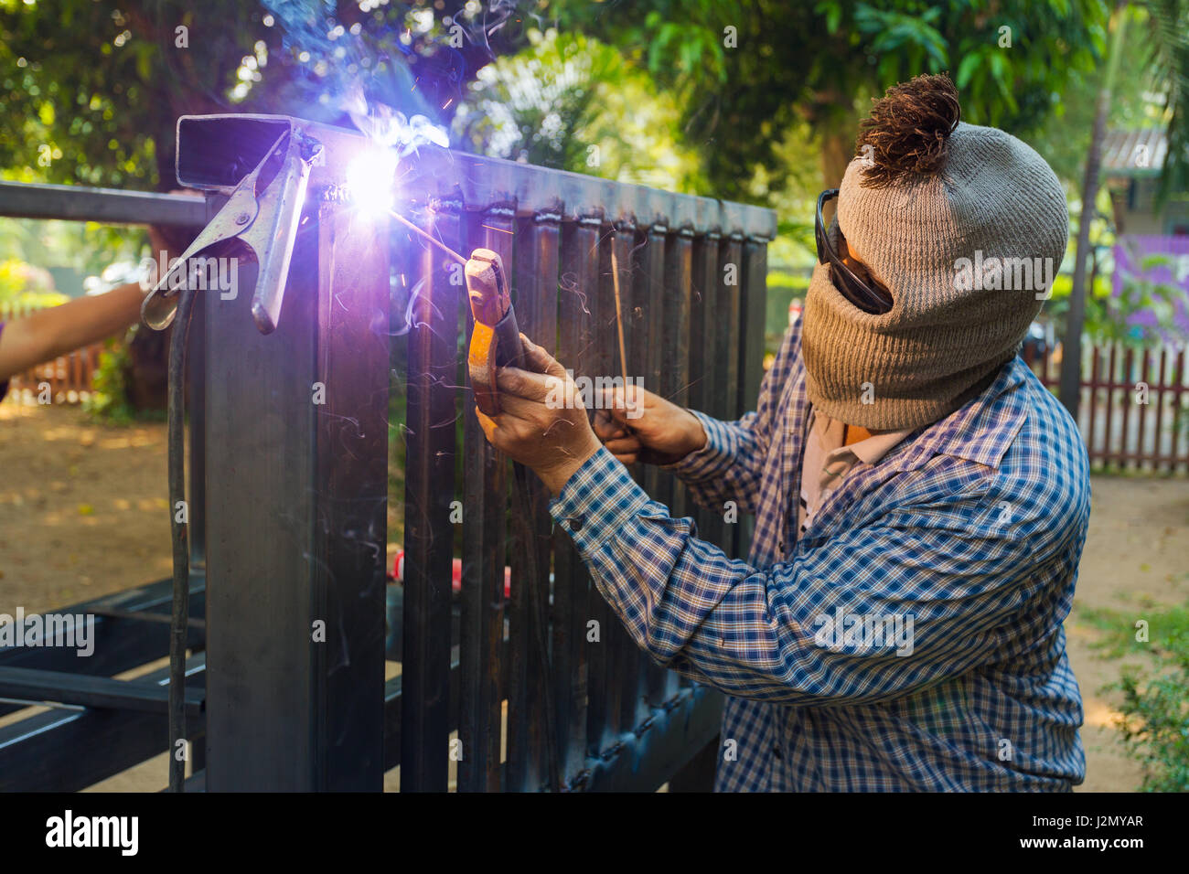 Unidentified hooded man welding steel platform, bright light and sparks, outdoor with blurr background trees Stock Photo